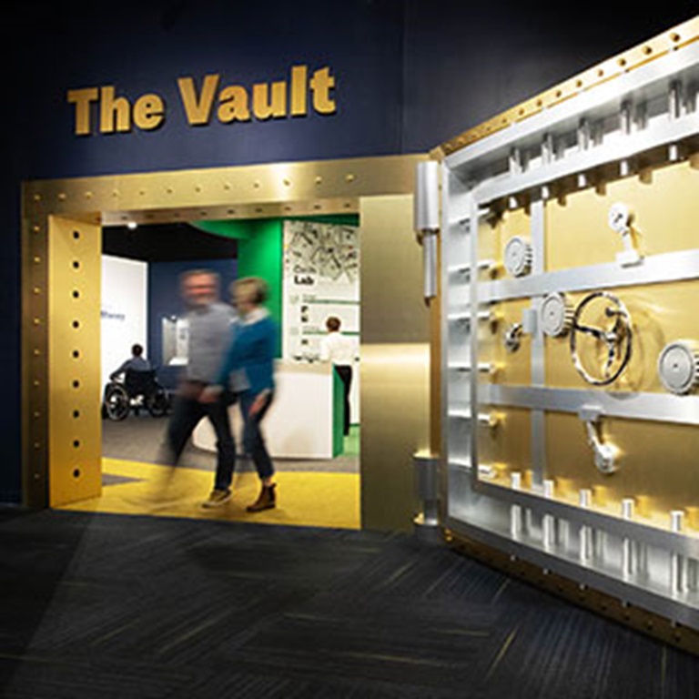 Two people walk past a large golden vault door.