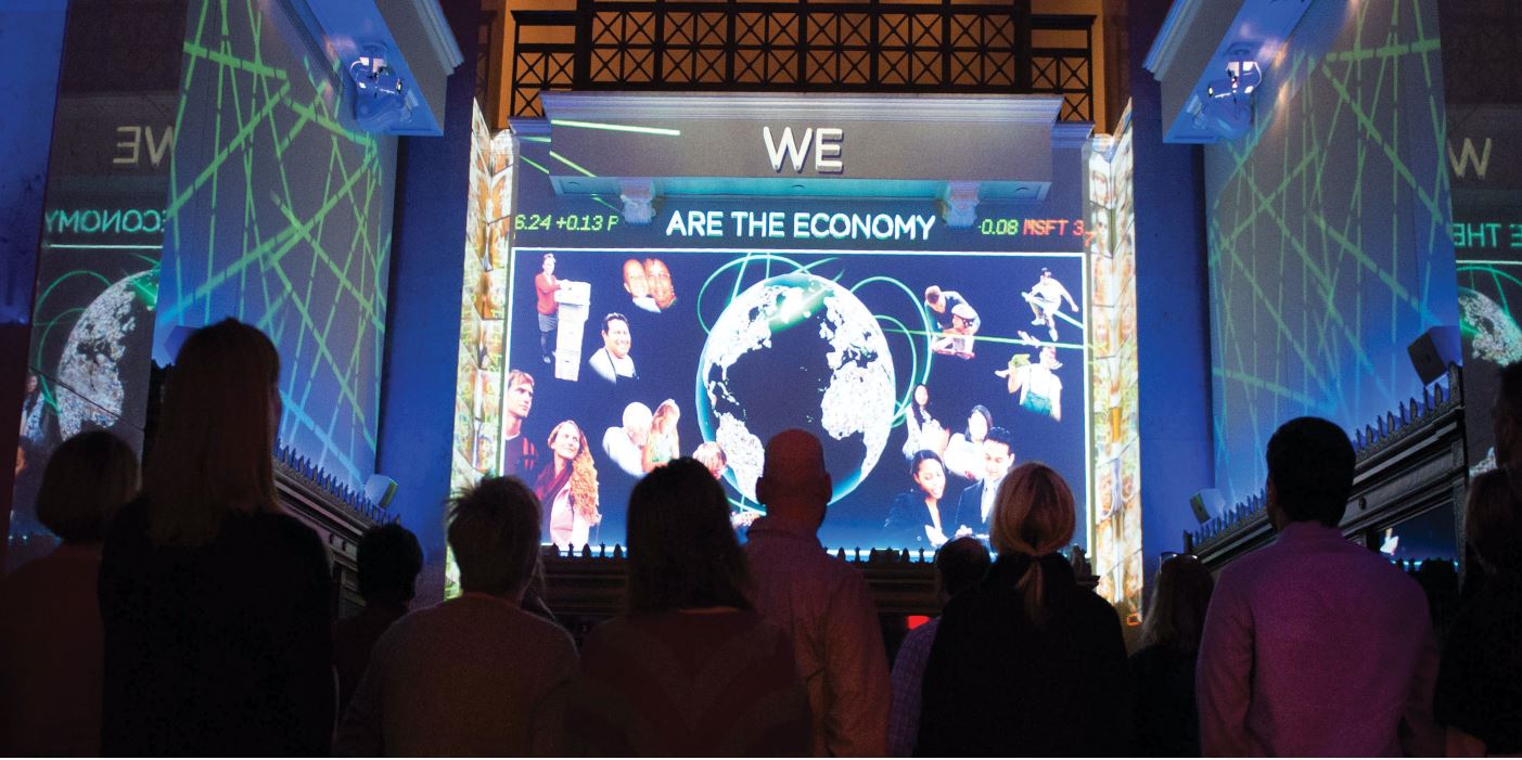 A large group of people in silhouette watch a digital display that reads 'we are the economy.'